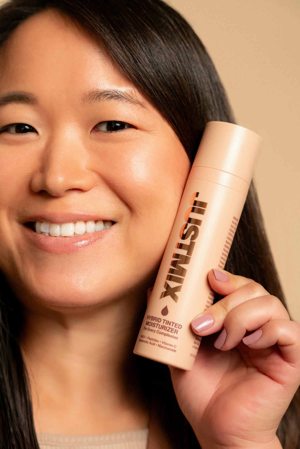 Woman smiling and holding JUSTMIX Hybrid Tinted Moisturizer beside her face