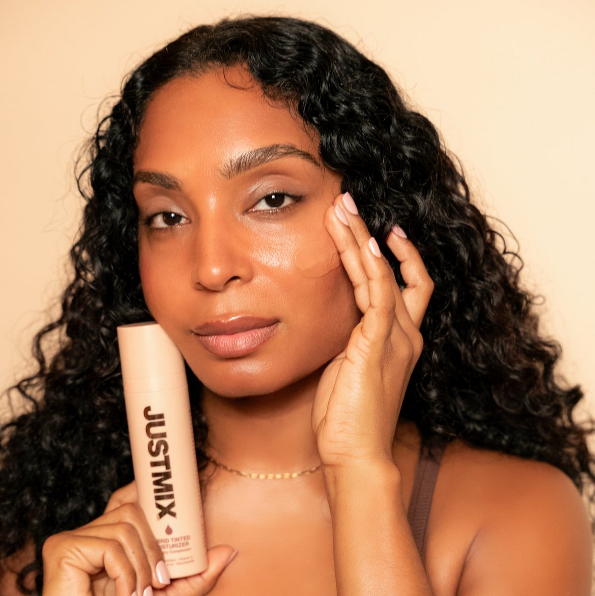 Woman holding JUSTMIX Hybrid Tinted Moisturizer, with the tint blended on her cheek
